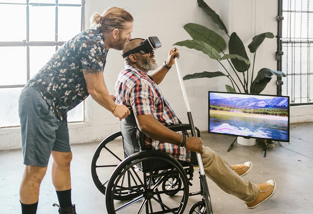 A man is holding the back of a wheelchair. The is another man sitting in the wheelchair wearing a VR set over his face holding an oar. In the background there is a TV with a POV of kakaying in a mountain lake.